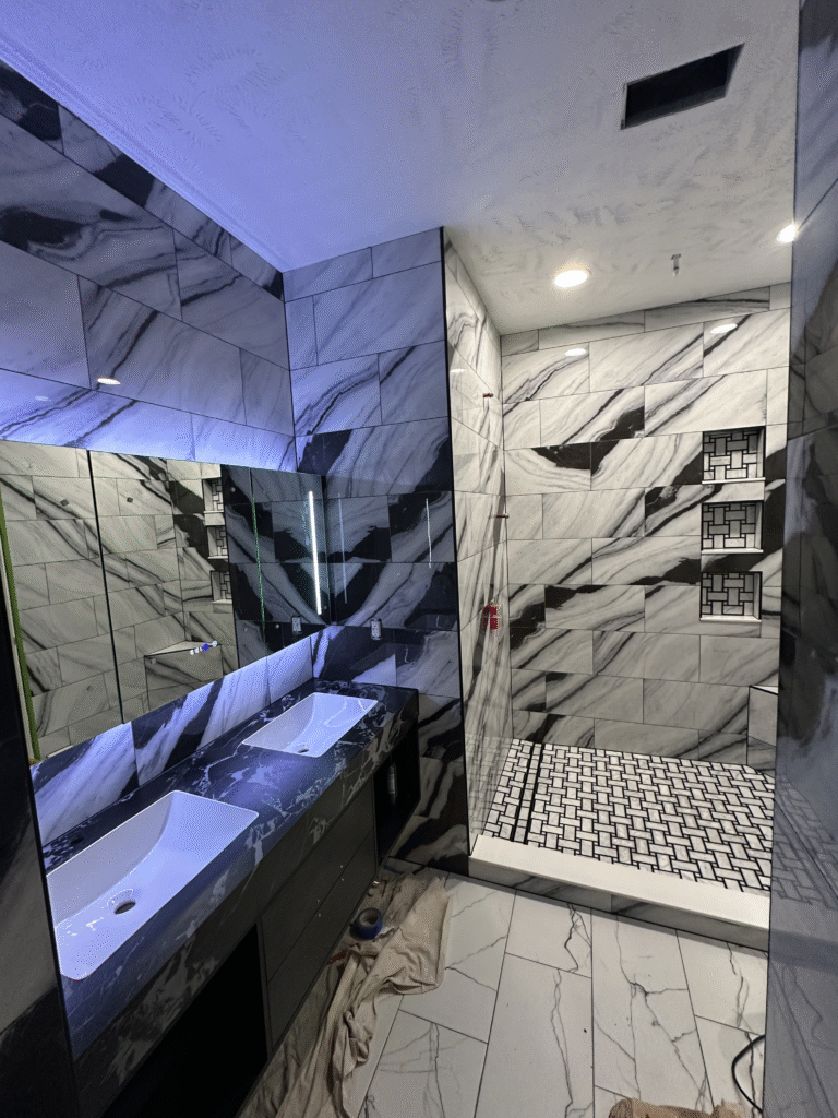 Completed luxury bathroom remodel in Arlington VA featuring double vanity, modern lighting, and marble tile shower with niche shelving.